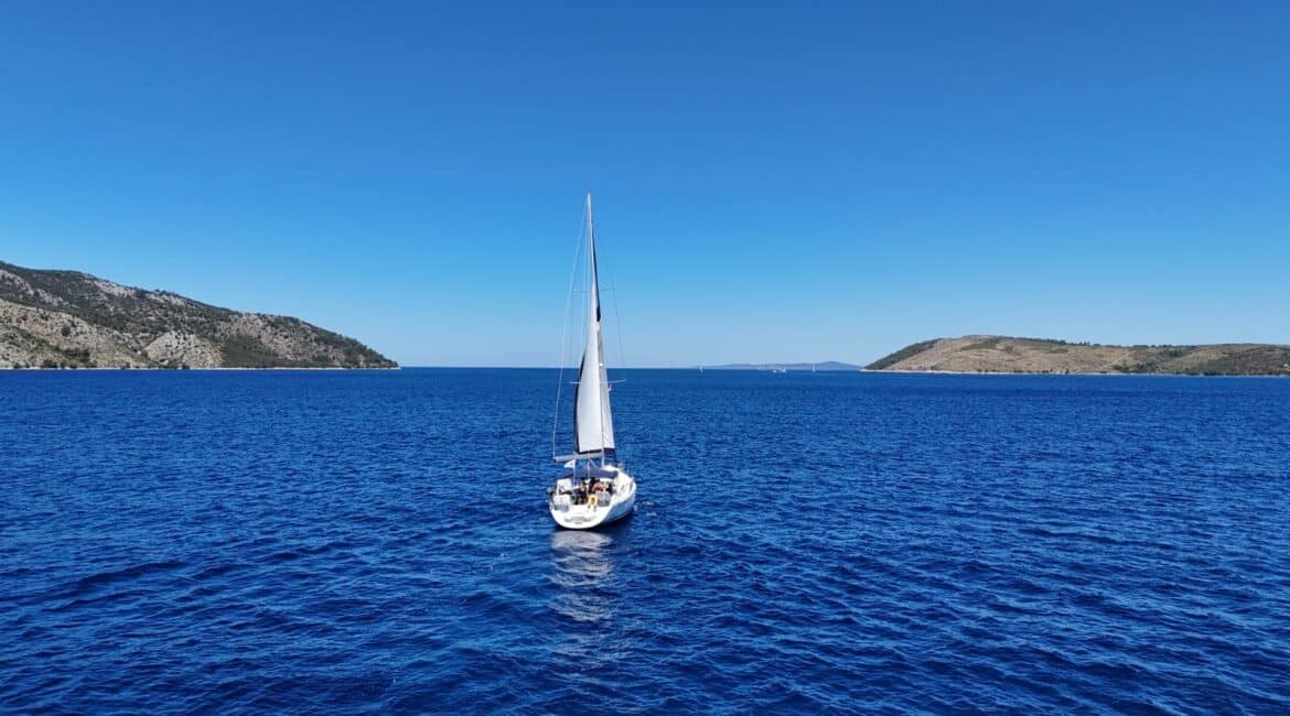 Sustainable sailing, A close-up of a sailor picking up a piece of plastic from a pristine white pebble beach with a sailing yacht anchored in the background.
